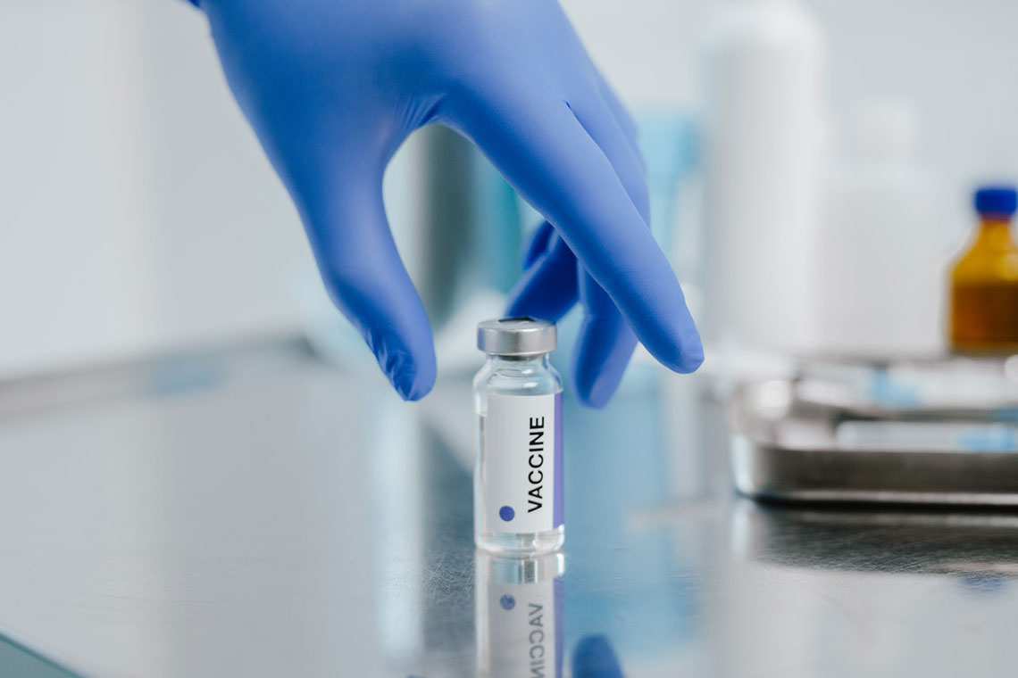 A hand in a blue latex glove reaches for a vial of a vaccine on a metal table